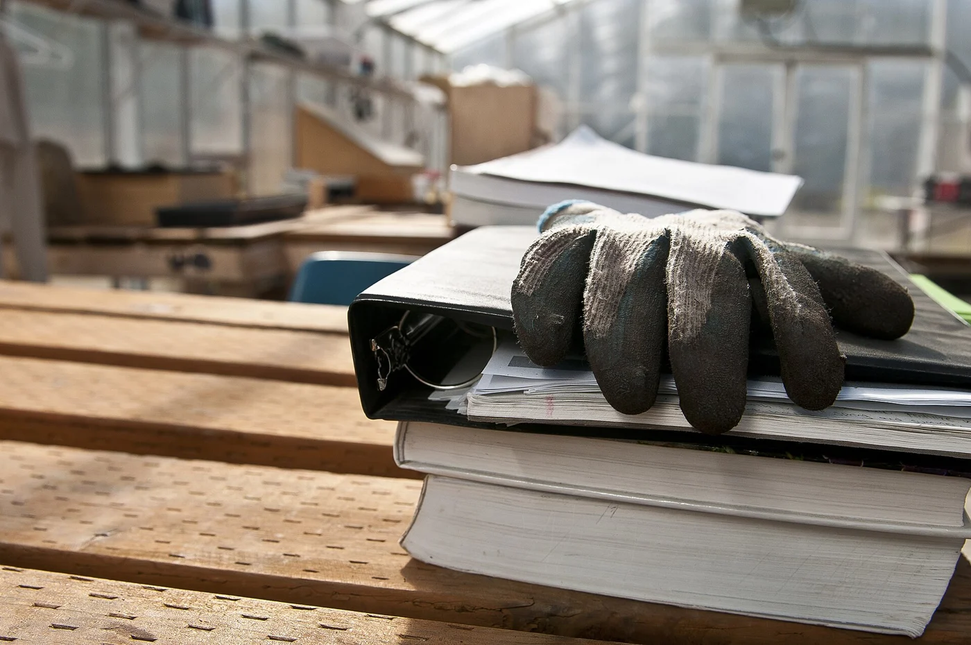 A gardening glove rests on an open folder and books on a wooden bench inside a greenhouse.