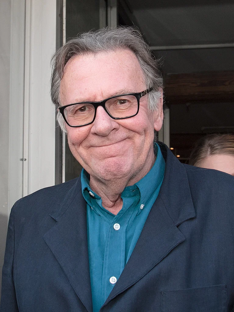 Tom Wilkinson wearing black-framed glasses and a jacket over a blue shirt.
