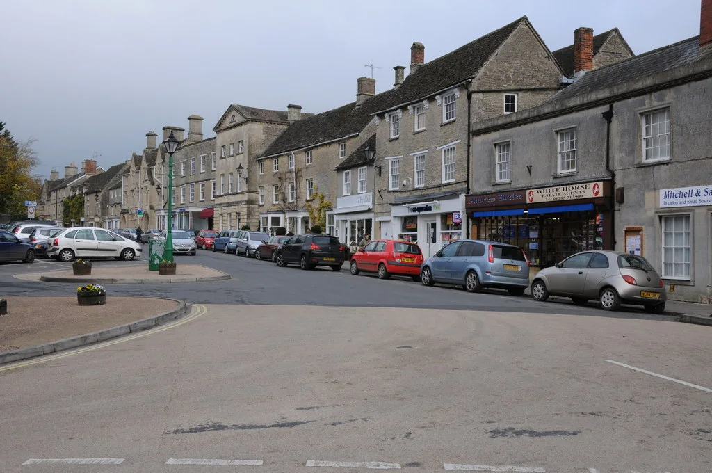 Stone-fronted shops and houses along High Street in Fairford, with cars parked beside the pavement.