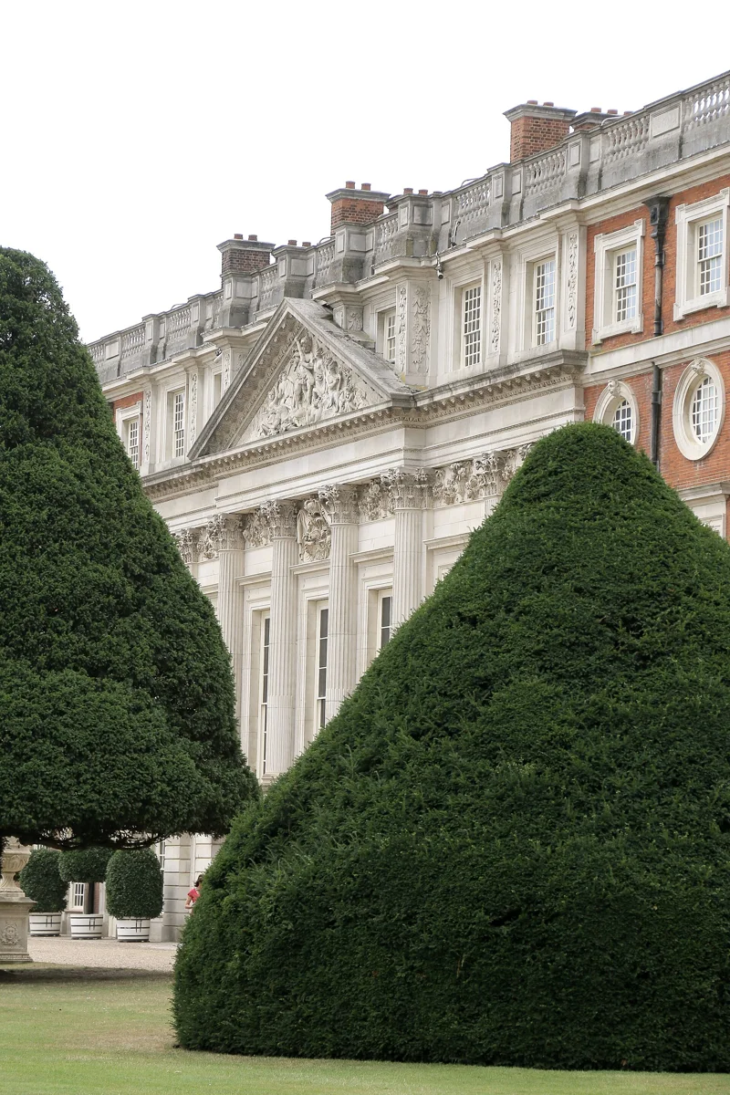 A pale stone facade of Hampton Court Palace behind tall clipped evergreen trees.