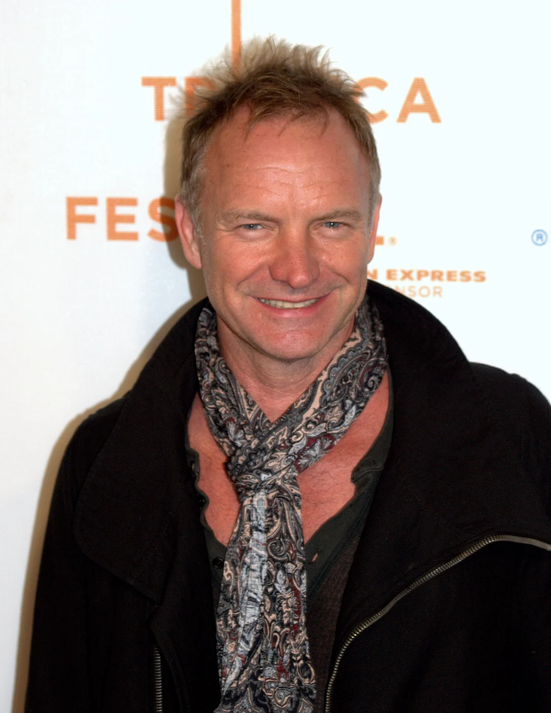 Sting wearing a dark coat and patterned scarf in front of a Tribeca Film Festival backdrop.